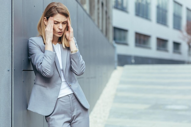 Une femme d'affaires près d'un immeuble de bureaux à l'extérieur a de graves maux de tête fatigués et déprimés, tenant les mains derrière la tête