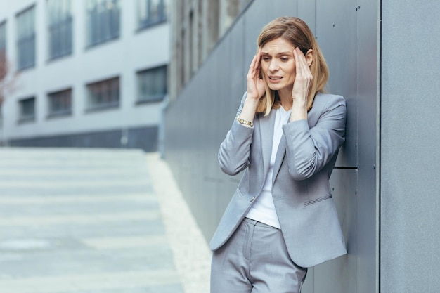 Une femme d'affaires près d'un immeuble de bureaux à l'extérieur a de graves maux de tête fatigués et déprimés, tenant les mains derrière la tête