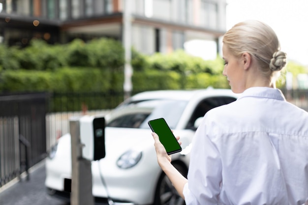 Une femme d'affaires progressiste en costume regarde l'état de la batterie de la voiture EV à partir d'un téléphone simulé