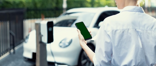 Une femme d'affaires progressiste en costume regarde l'état de la batterie de la voiture EV à partir d'un téléphone simulé