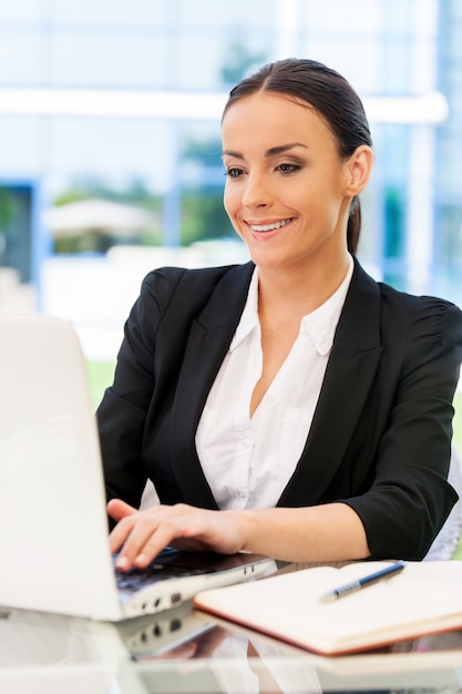 Femme d'affaires prospère. Belle jeune femme en tenue de soirée travaillant sur ordinateur portable et souriante assise à la table à l'extérieur