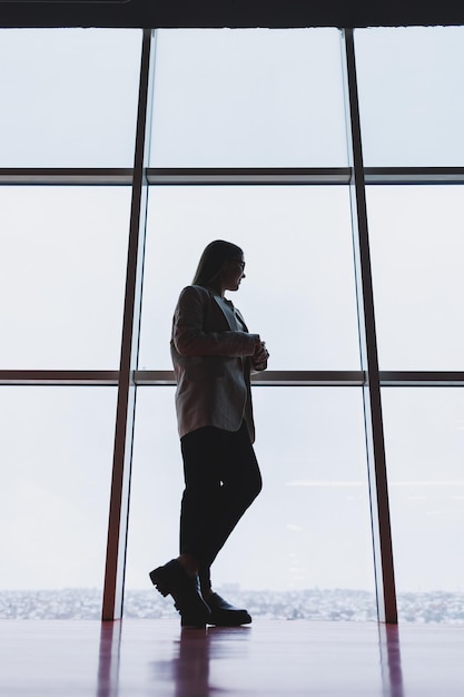 Une femme d'affaires prospère en pantalon et veste se tient debout et regarde par l'immense fenêtre panoramique du gratte-ciel du centre d'affaires