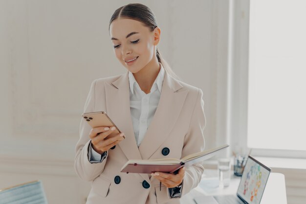 Femme d'affaires prospère regardant l'écran du smartphone avec le bloc-notes à la main, lisant les dernières nouvelles ou vérifiant les e-mails. Employée souriante en costume formel beige travaillant dans un bureau moderne et léger