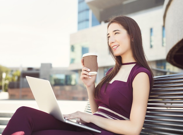 Femme d'affaires réfléchie travaillant sur ordinateur portable. Agent d'assurance caucasien vérifiant ses e-mails sur ordinateur, buvant du café, assis sur le banc à l'extérieur près d'un centre de bureaux moderne