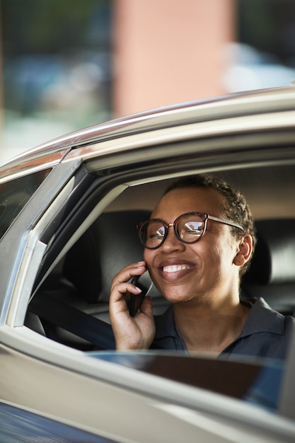 Femme d'affaires réussie dans des lunettes parlant au téléphone portable et riant pendant son voyage en voiture