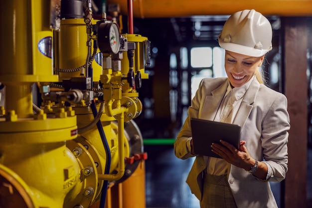 Femme d'affaires réussie dédiée blonde dans le contrôle de l'usure formelle sur les machines et tenant la tablette en position debout dans la centrale électrique.