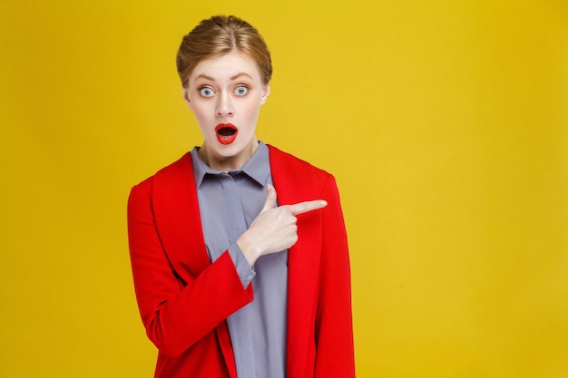 Femme d'affaires rousse choquée en costume rouge montrant à l'espace de copie. Studio shot, isolé sur fond jaune