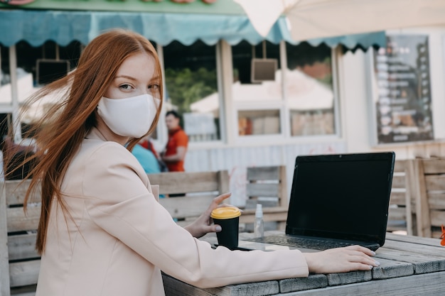 Femme d'affaires rousse portant un masque facial pour la protection contre les virus travaillant sur l'ordinateur portable à l'extérieur. Jeune femme d'affaires travaillant sur ordinateur portable à l'extérieur.