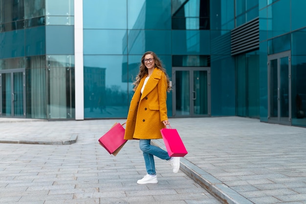 femme affaires, à, sacs provisions, habillé, manteau jaune, marche, dehors, corporatif, bâtiment, fond, caucasien, femme, personne affaires, immeuble bureau, élégant, femme affaires, saison, vente, pleine longueur