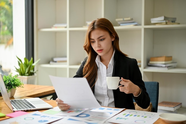 Une femme d'affaires se concentre sur l'examen des rapports financiers et sirote un café à son bureau