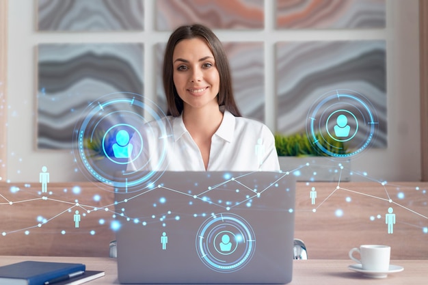 Femme d'affaires séduisante en chemise blanche sur le lieu de travail travaillant avec un ordinateur portable pour embaucher de nouveaux employés pour le conseil en affaires internationales Icônes d'hologramme de médias sociaux RH sur fond de bureau