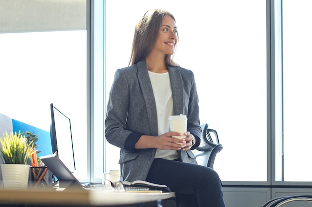 Une femme d'affaires séduisante tient une boisson chaude assise au bureau.