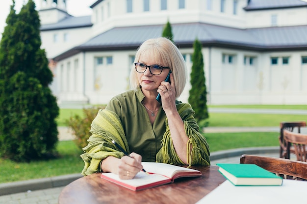 Femme d'affaires senior belle assise sur une chaise et prendre des notes dans un ordinateur portable, avec un téléphone et un ordinateur portable dans la cour