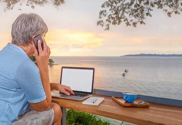 Femme d'affaires senior avec ordinateur portable et tasse de café à table dans un café avec vue sur la mer parle par téléphone