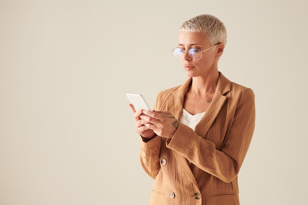 Femme d'affaires sérieuse aux cheveux blonds dans des verres ronds debout sur fond blanc et à l'aide de smart