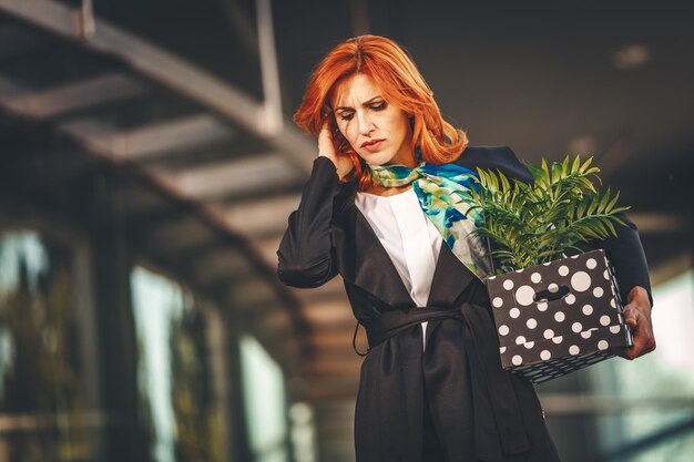 Une femme d'affaires sérieuse et pensive se promène dans le quartier des bureaux et parle sur un smartphone avec une boîte pleine de ses effets personnels du bureau juste après son licenciement.