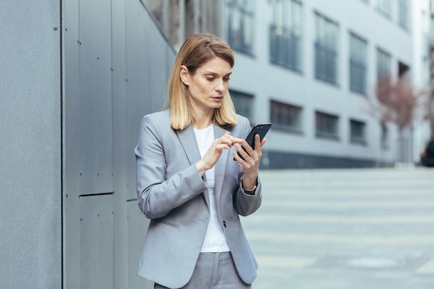 Une femme d'affaires sérieuse et pensive utilise le téléphone à l'extérieur du bureau en regardant l'écran du smartphone