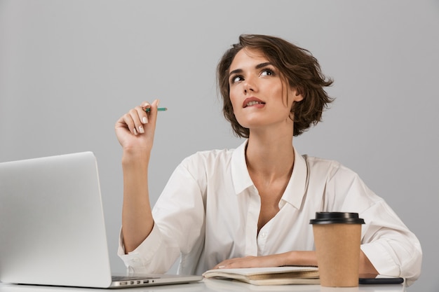 Femme d'affaires sérieuse posant isolé sur mur gris assis à la table à l'aide d'un ordinateur portable