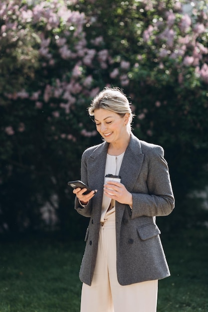 Femme d'affaires souriante blonde mature marchant dans le parc en buvant du café et en parlant au téléphone au printemps sur un fond de fleurs lilas