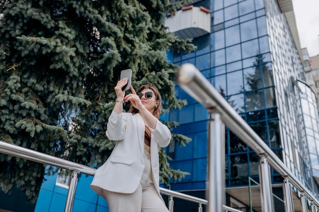 Femme d'affaires souriante en costume blanc et lunettes de soleil faisant selfie à l'aide de téléphone pendant la pause debout près d'un immeuble de bureaux moderne