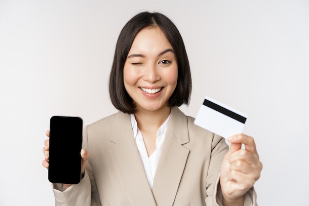 Femme d'affaires souriante en costume montrant l'écran du téléphone portable et l'application sur l'écran du smartphone du téléphone portable debout sur fond blanc
