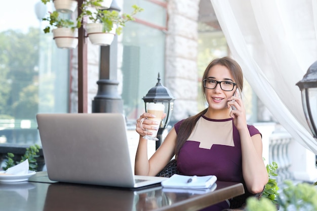 Femme d'affaires souriante parlant au téléphone, buvant du café et travaillant sur un ordinateur portable, assise au café. Travailleur indépendant, concept de mode de vie de jeune fille occupée, espace de copie