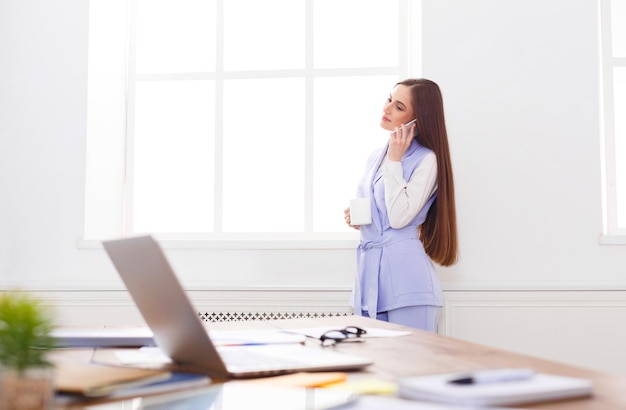 Femme d'affaires souriante parlant par téléphone près de la fenêtre sur le lieu de travail du bureau, espace de copie. Communication, technologie, concept de réussite