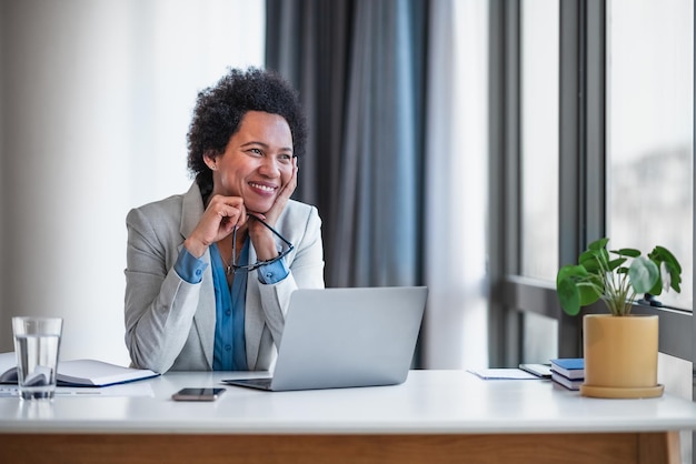 Femme d'affaires souriante et réfléchie regardant loin tout en utilisant un ordinateur portable au bureau