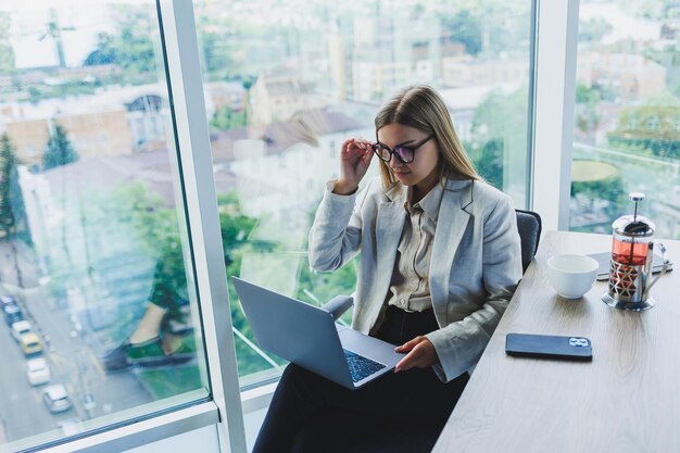 Femme d'affaires souriante regardant un ordinateur portable tout en travaillant au bureau Le concept d'une femme moderne qui réussit L'idée des affaires et de la vie d'un entrepreneur Jeune femme à table avec un ordinateur portable ouvert