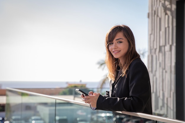 Une femme d'affaires souriante répond aux e-mails sur son smartphone depuis le balcon de son bureau