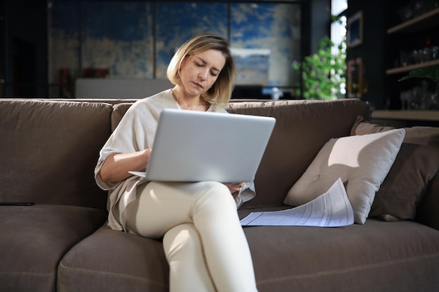 Une femme d'affaires souriante s'assoit sur un canapé en regardant un webinaire sur un ordinateur portable. Heureuse femme d'âge moyen étudie sur un cours à distance en ligne.