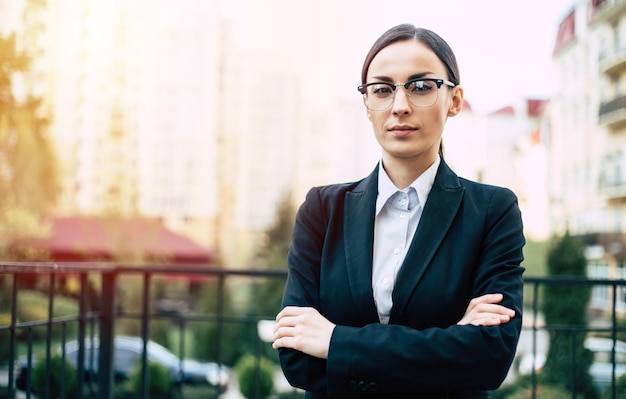 Femme d'affaires de succès moderne en lunettes et costume noir avec les bras croisés regardant la caméra sur fond de ville urbaine