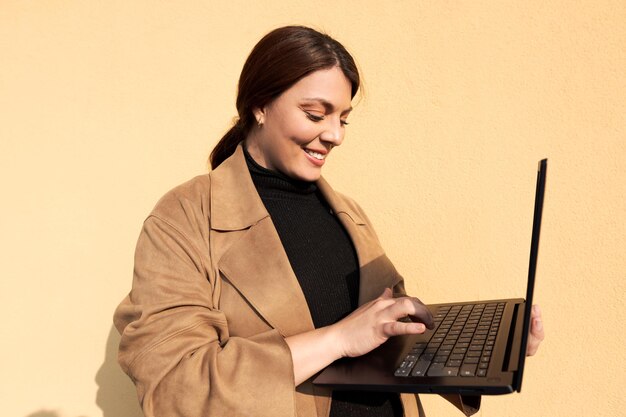 Photo femme d'affaires tapant sur son ordinateur portable tout en souriant portant un manteau marron et les cheveux attachés en arrière