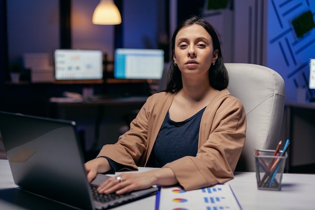 Femme d'affaires tenant la main sur le clavier d'un ordinateur portable en regardant la caméra. Femme intelligente assise sur son lieu de travail pendant les heures tardives de la nuit faisant son travail.