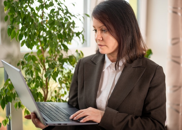 Femme d'affaires tenant un ordinateur portable. femme adulte portant un costume tenant un ordinateur portable argenté au bureau sur fond de fenêtre. Femme adulte tenant un ordinateur portable et regardant l'écran