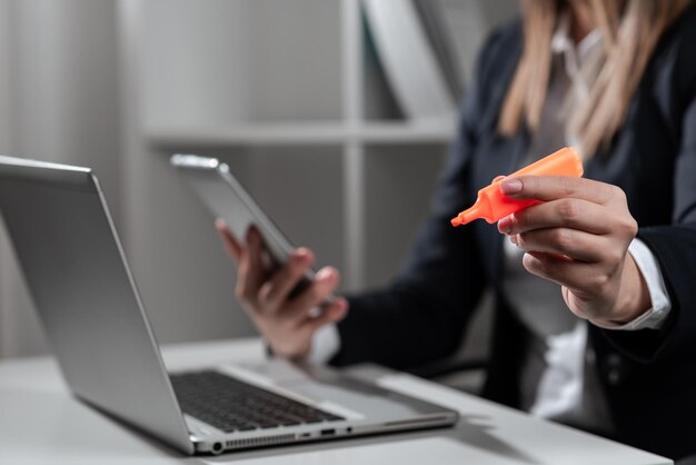 Femme d'affaires tenant un téléphone et pointant avec un marqueur sur un message important sur le bureau avec un ordinateur portable en costume présentant des informations cruciales