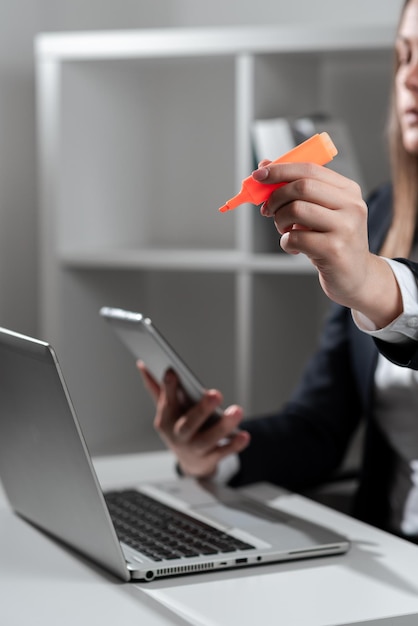 Femme d'affaires tenant un téléphone et pointant avec un marqueur sur un message important sur le bureau avec un ordinateur portable en costume présentant des informations cruciales