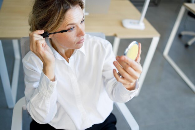 Femme d'affaires en tenue de soirée avec un miroir à la main utilise du mascara