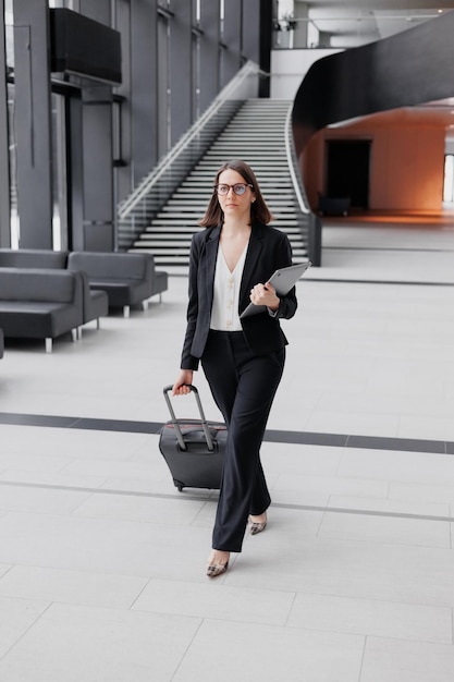 Femme d'affaires de tourisme d'affaires en costume formel avec un smartphone dans un centre d'affaires ou un aéroport avec une valise part en voyage d'affaires