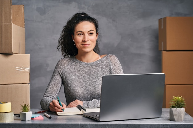 Femme d'affaires travaillant à domicile avec un ordinateur portable et un bloc-notes préparant des boîtes avec des produits à livrer