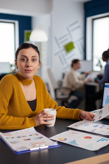 Femme d'affaires travaillant avec des graphiques d'entreprise regardant la caméra tenant une tasse de café