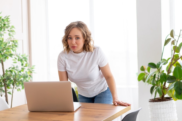 Femme d'affaires utilisant un ordinateur portable debout près du bureau intérieur de bureau blanc avec plante d'intérieur regardant la caméra Gens d'affaires Homme d'affaires en ligne, jeune et prospère chemise blanche et jeans Dresed