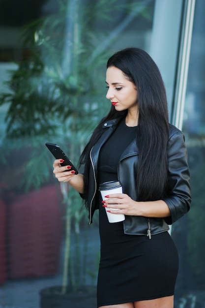 Femme d'affaires utilisant un smartphone numérique et buvant du café près de l'immeuble de bureaux