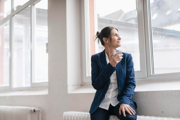 Femme d'affaires avec un verre d'eau assis sur un radiateur dans un loft regardant par la fenêtre