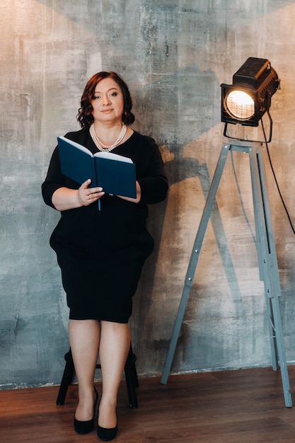 Une femme d'affaires vêtue d'une robe perlée noire se tient contre le mur avec un livre dans ses mains.