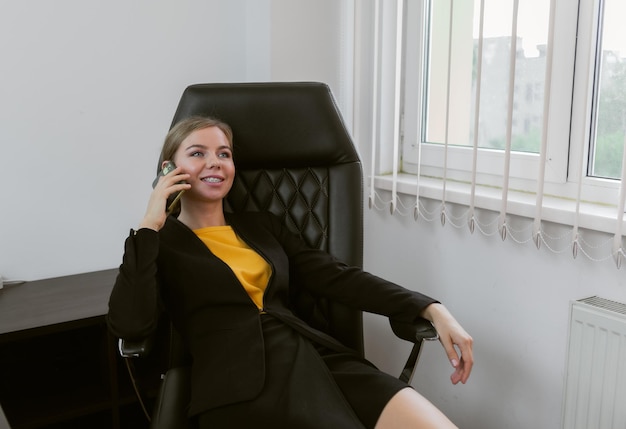 Femme d'affaires vêtue de vêtements confiants parlant au téléphone tout en étant assise dans la chaise du patron dans son bureau