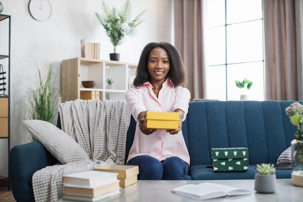 Femme africaine assise sur un canapé avec une boîte-cadeau jaune dans les mains