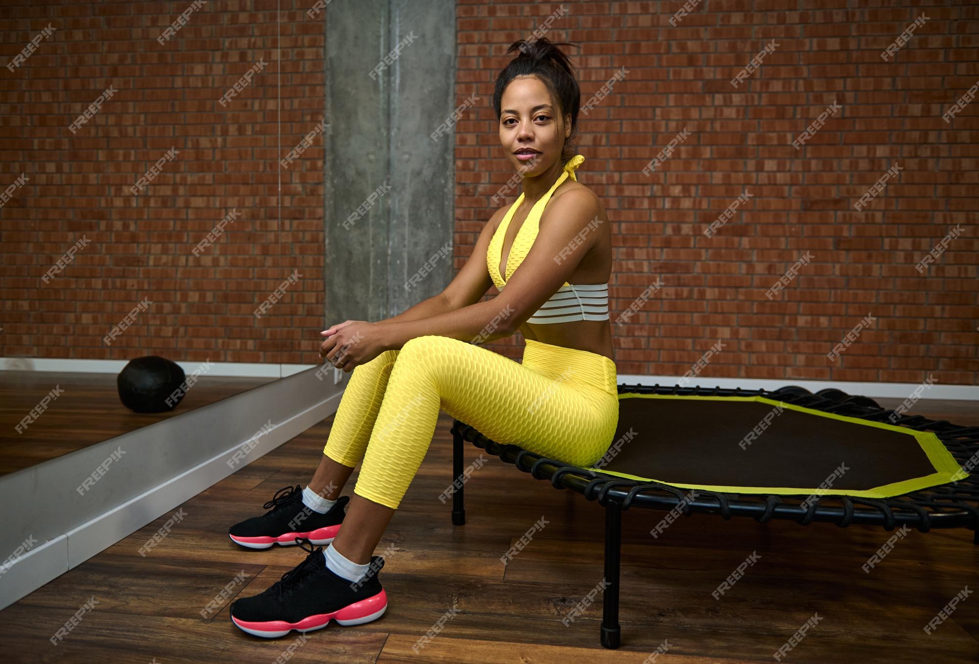 Femme Africaine Au Corps Mince Et Au Ventre Plat En Survêtement Jaune Vif,  Assise Sur Un Trampoline En Posture De Lotus Et Souriante En Regardant La  Caméra. Fitness, Sport, Méditation, Yoga, Concept De Mode De Vie Actif