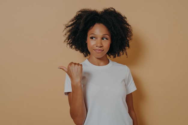 Une femme africaine aux cheveux assez bouclés pointe le pouce de côté, recommande une promotion et fait de la publicité pour un produit avec une expression heureuse, suggère de visiter cet endroit tout en posant sur fond beige