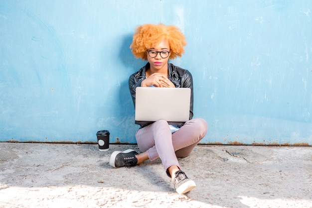 Photo femme africaine aux cheveux bouclés travaillant avec un ordinateur portable assis sur le fond bleu du mur